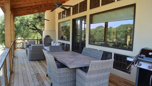 Un patio techado al aire libre con mobiliario de mimbre, una parrilla y amplias ventanas con vistas a los árboles.