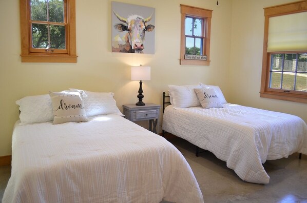 Un dormitorio con dos camas, cada una con ropa blanca y almohadas, una mesita de noche con lámpara, y una obra de arte en la pared que representa una vaca.