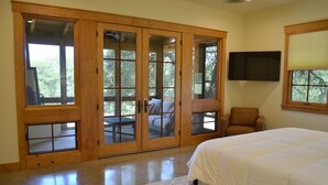Un dormitorio con una cama grande, una puerta corredera de madera que da a un balcón, un televisor montado y una silla.