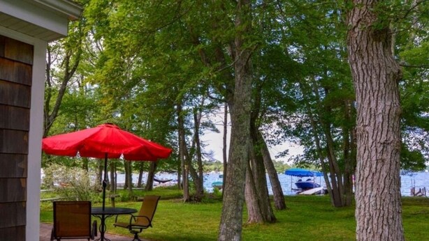 An outdoor patio with a table and chairs under a red umbrella, surrounded by trees and a view of a lake. - Charming cottage on the water with beautiful views (Lakeville)