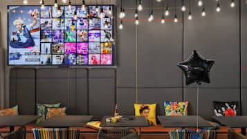 A modern lounge area with a large TV displaying various images, multiple seating arrangements, and hanging light bulbs.
