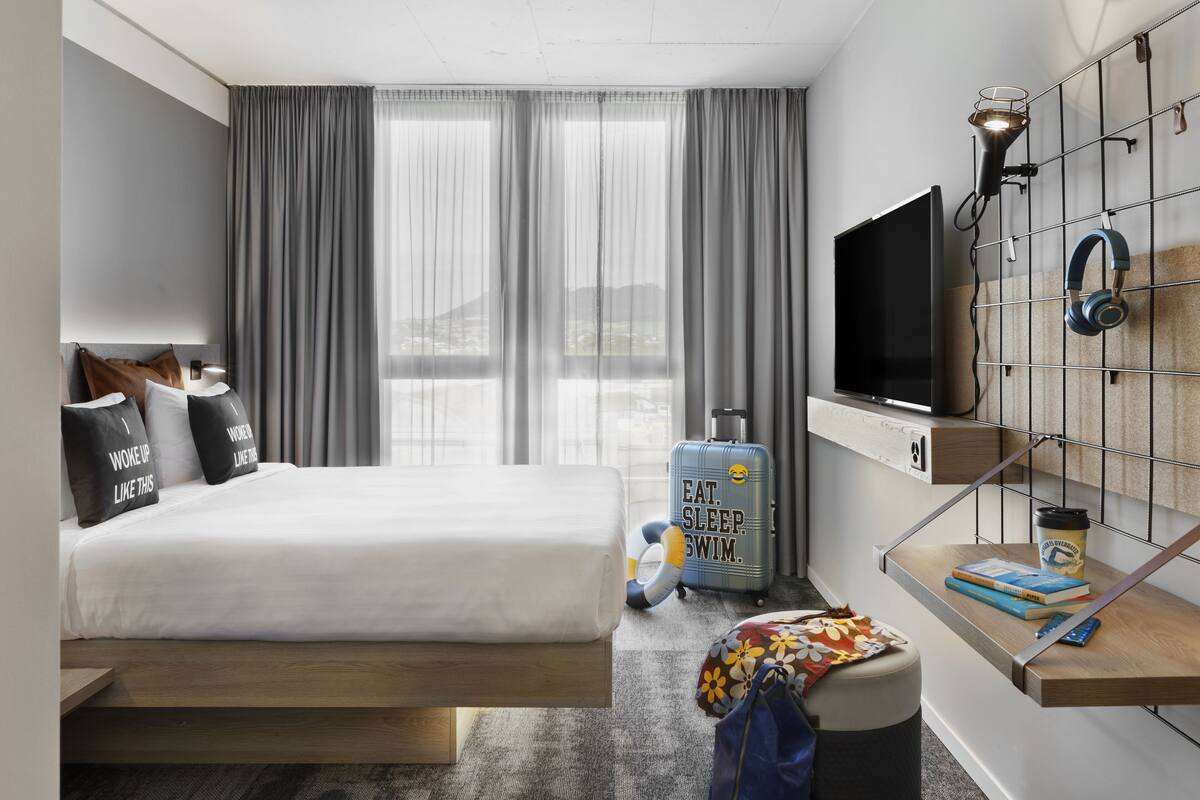 Habitación de hotel con cama, televisor, maleta y estante en la pared con auriculares y taza.