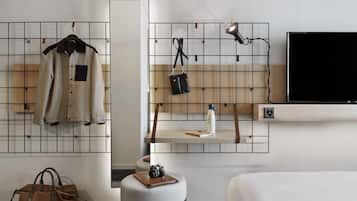 A hotel room with a bed, a hanging coat rack, a small table with a water bottle, a camera, and a brown leather bag.