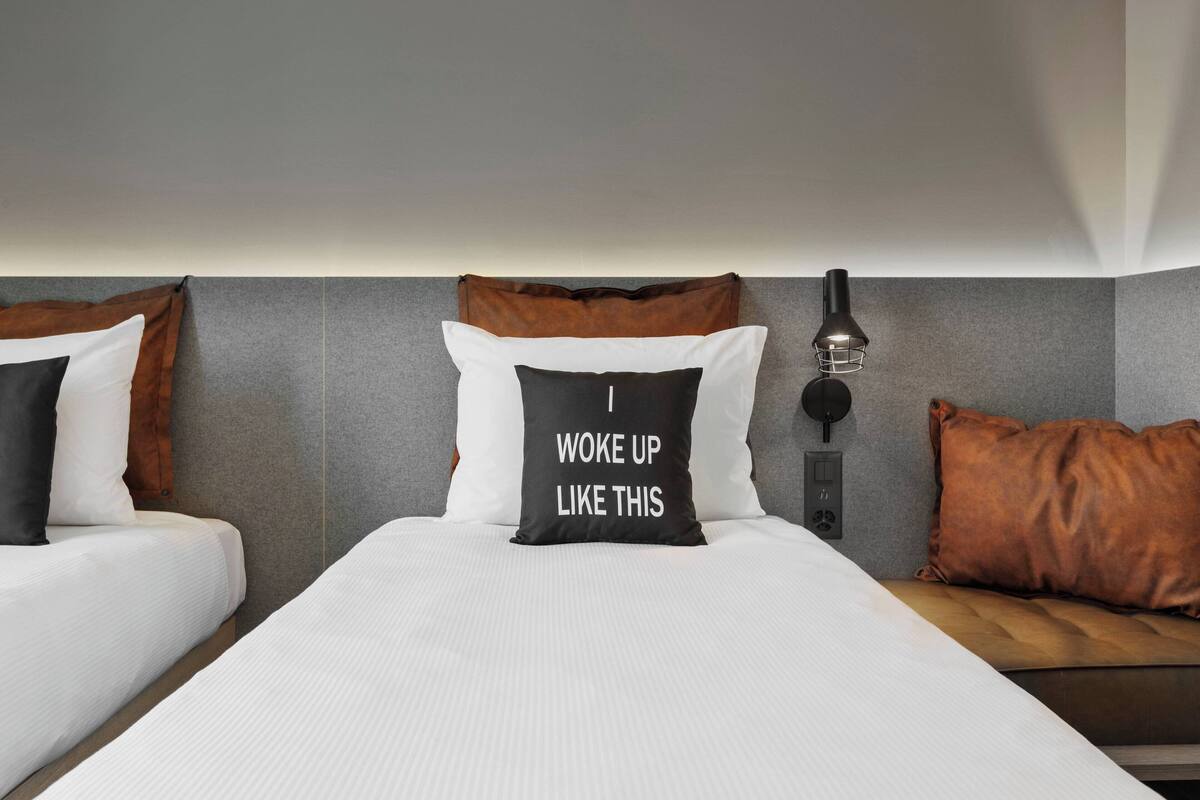 Habitación de hotel con dos camas, una mesita de noche, una lámpara y un aplique de pared.