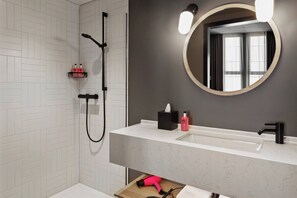 Un baño moderno con ducha de azulejos blancos, un espejo grande y un lavabo blanco con grifo negro.