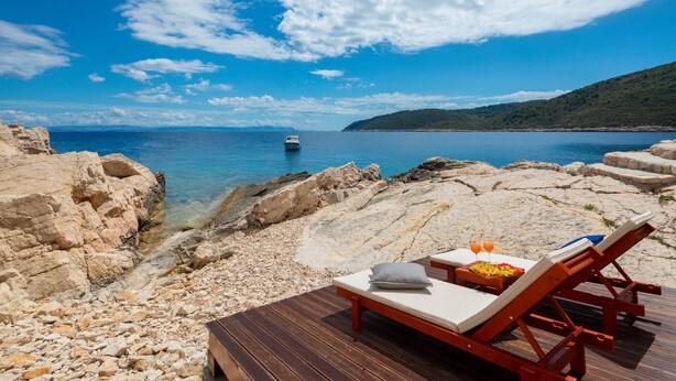 A beach with two wooden lounge chairs, a table with fruit and drinks, and a boat in the distance. - Luxury Villa Lighthouse Beyond (Vis)