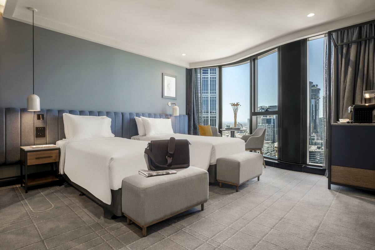 A hotel room with a large bed, a desk, a chair, and a view of the city through large windows.