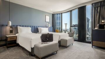 Habitación de hotel con una cama grande, un escritorio, una silla y vistas de la ciudad a través de amplios ventanales.