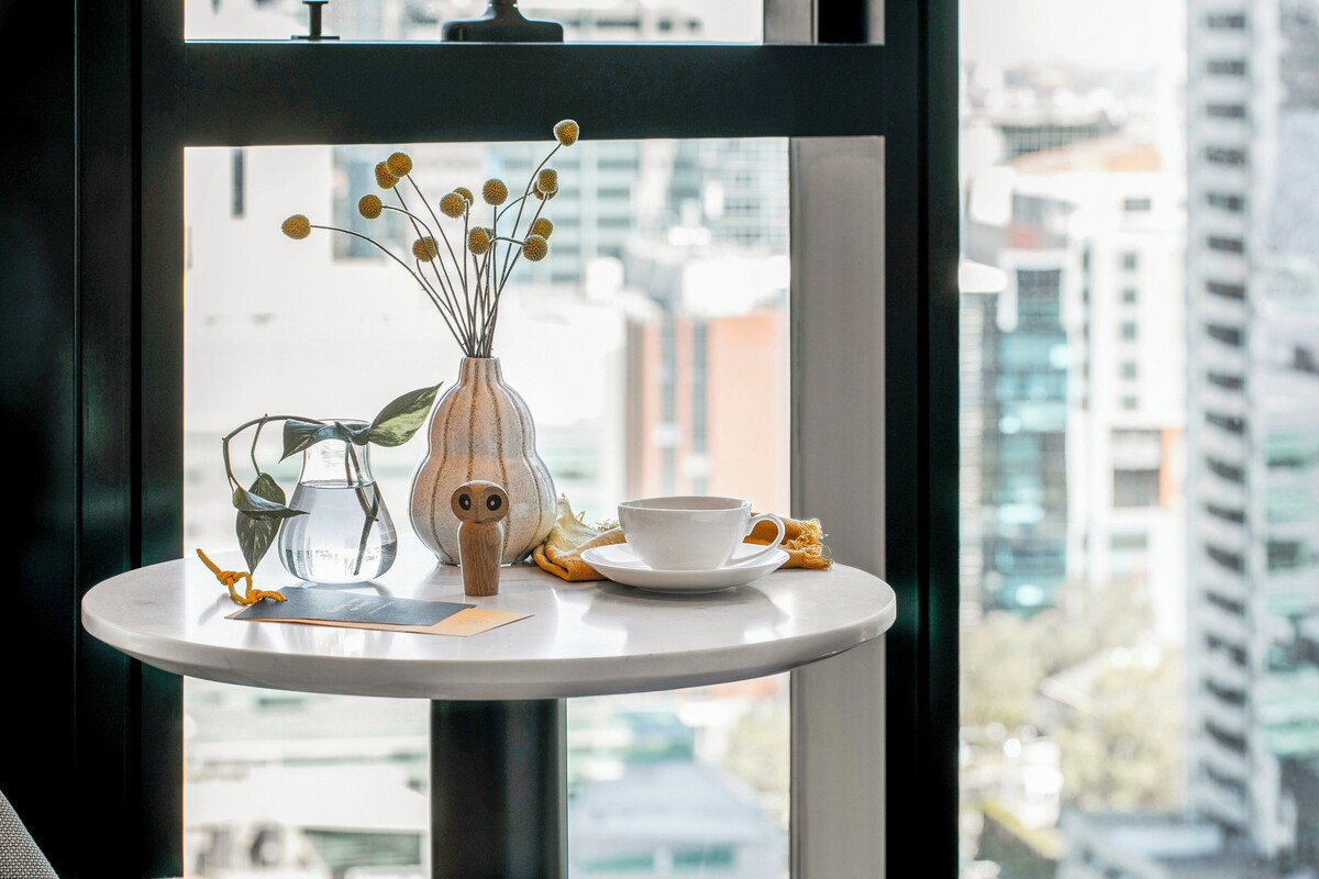 Una pequeña mesa redonda con una taza y platillo blancos, un florero con flores secas y una figurita de lechuza de madera.