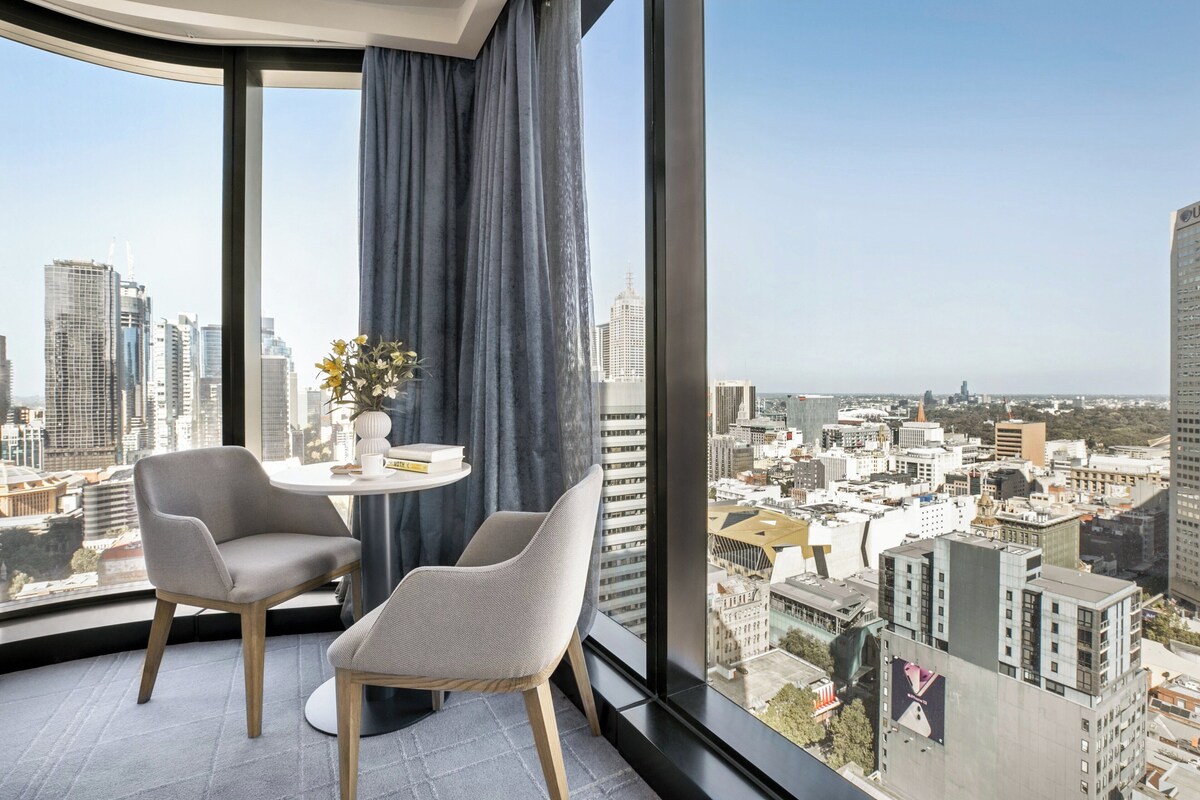 Habitación de hotel moderna con un ventanal grande que ofrece vistas a la ciudad, dos sillas, una mesita y un florero con flores.