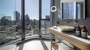 Un baño moderno con un ventanal grande, un espejo redondo, un lavabo sobre encimera y dos botellas de crema corporal.