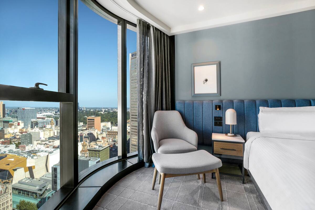 A hotel room with a curved window offering a city view, a bed, a chair, a bedside table with a lamp, and a framed picture on the wall.