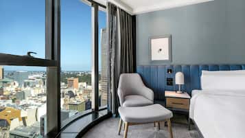 Habitación de hotel con ventanal curvo que ofrece vistas a la ciudad, cama, sillón, mesita de noche con lámpara y un cuadro en la pared.