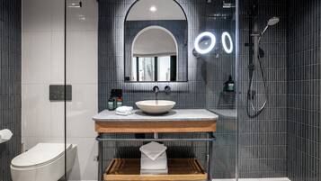 Un baño moderno con cabina de ducha de cristal, inodoro blanco, tocador de madera con lavabo y un espejo grande con vista a la ciudad.