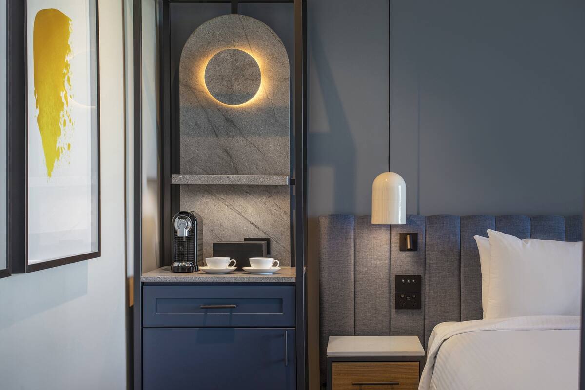 A modern hotel room with a bed, a nightstand, a coffee maker, and a wall-mounted circular light fixture.