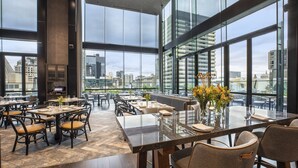 Un comedor amplio con techos altos y ventanas grandes que ofrecen vistas a la ciudad. Las mesas están dispuestas con sillas y centros de mesa.