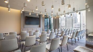 Un salón de conferencias con filas de sillas, una pantalla grande y lámparas colgantes.