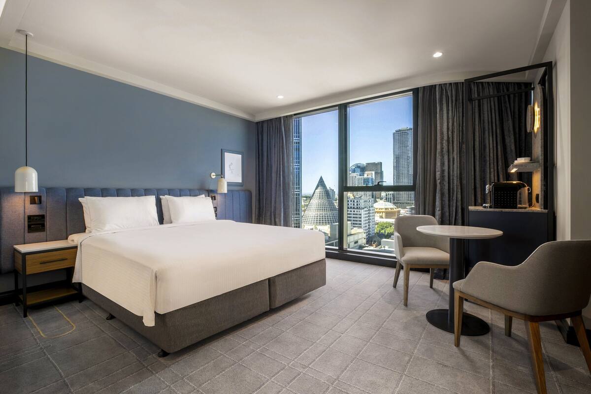 A modern hotel room with a large bed, a desk, a chair, and a view of the cityscape through large windows.