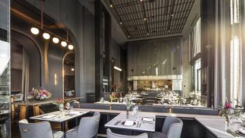 A modern restaurant with high ceilings, exposed beams, and a central bar area. Tables are set with white tablecloths and silverware.