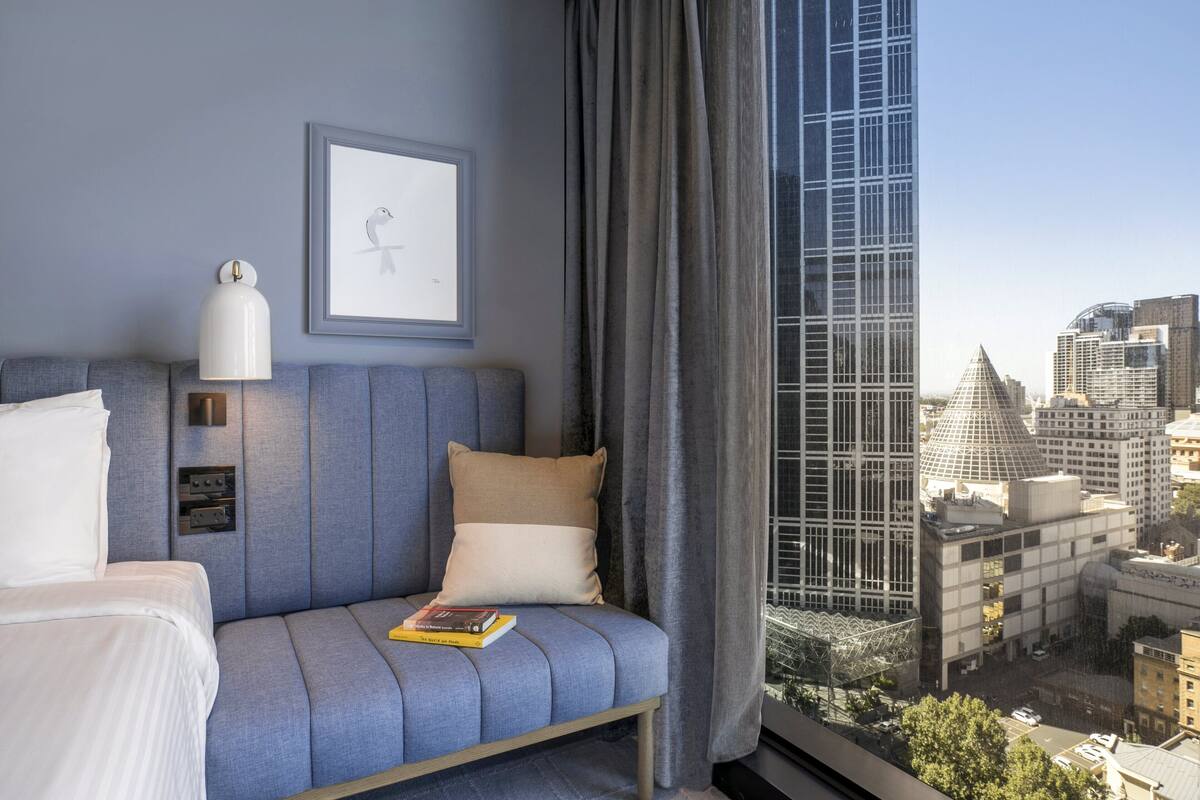 A modern hotel room with a large window, a city view, a sofa, a bedside table, and a framed picture on the wall.