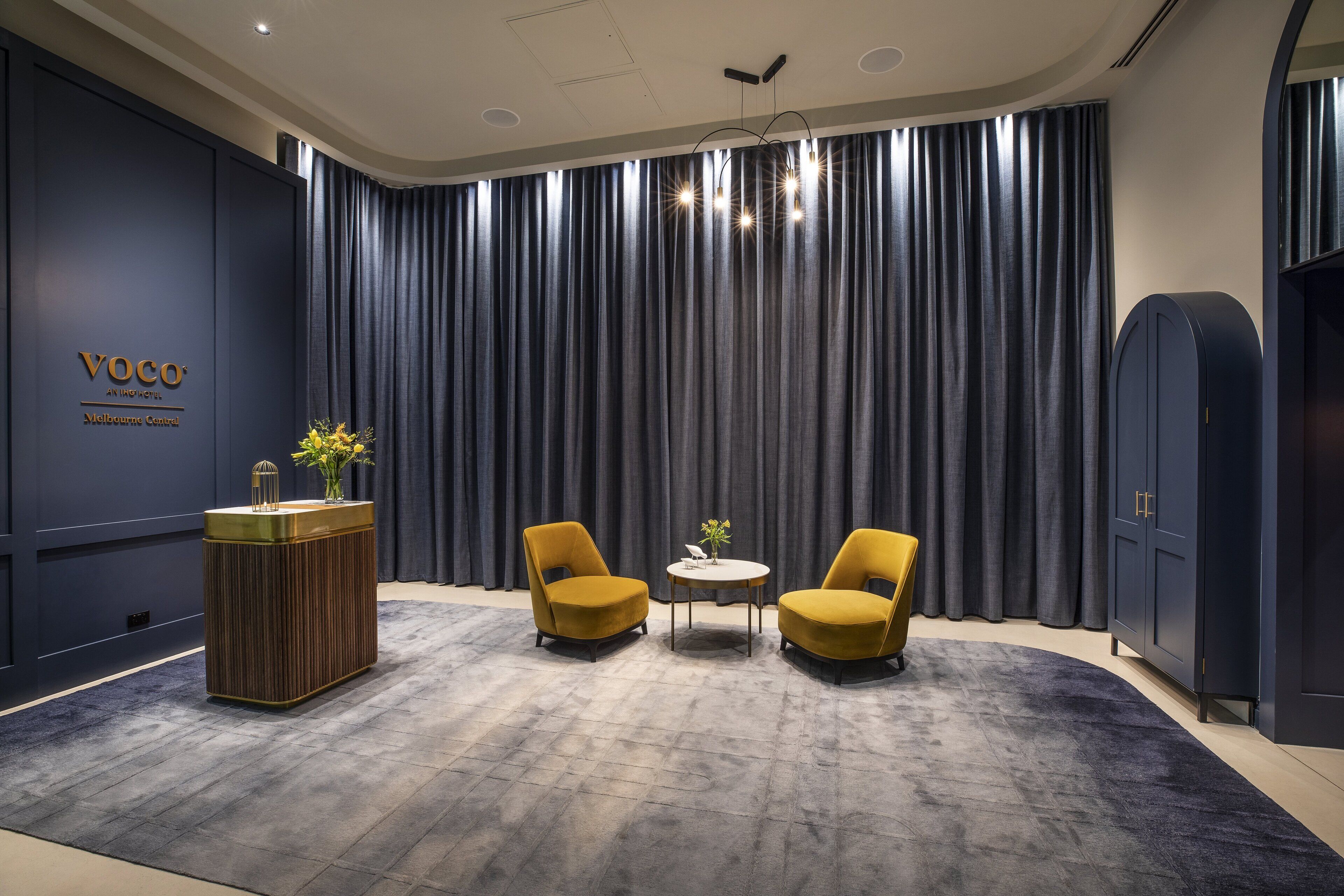 A modern hotel lobby with a dark blue wall, a reception desk, two yellow armchairs, a small round table, and a large curtain backdrop.