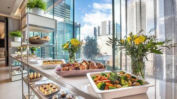 Un bufé con diversos platos, como pollo asado, verduras y postres, montado en una habitación de hotel moderna con ventanas amplias y vistas a la ciudad.