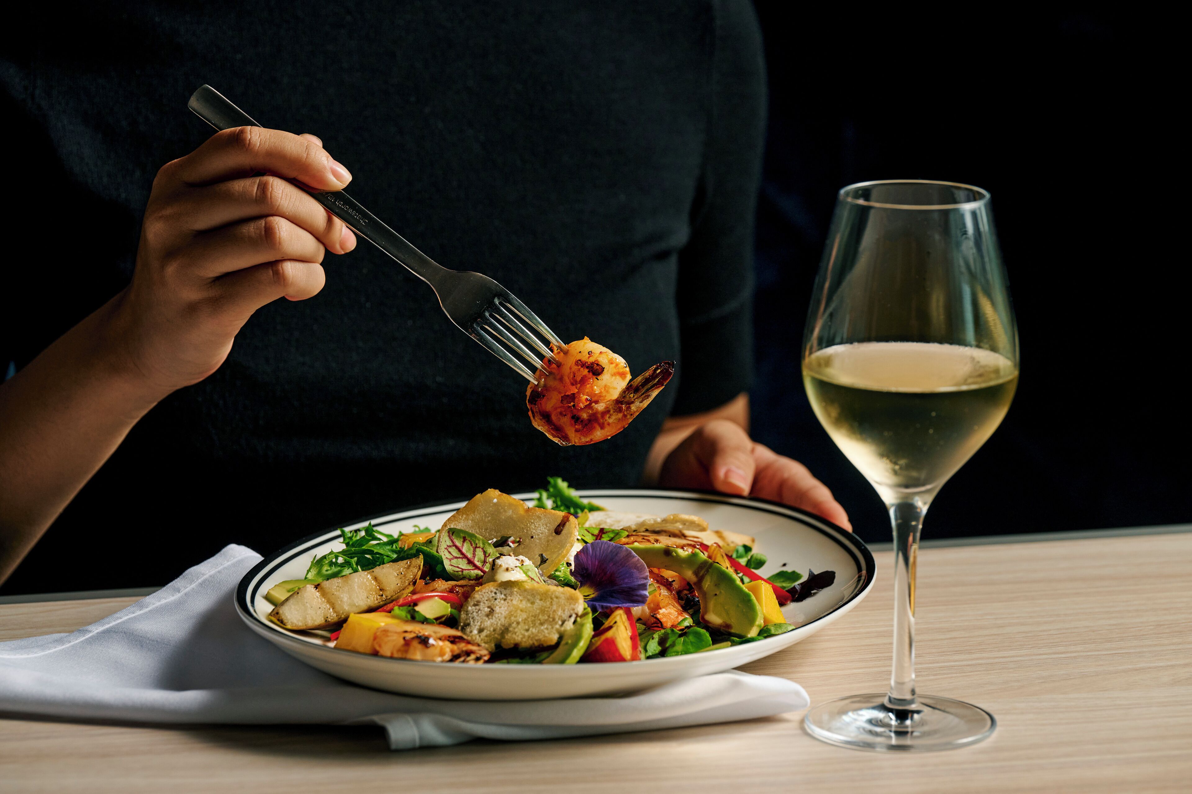 A person holding a fork with food, a glass of white wine, and a plate of salad on a table.