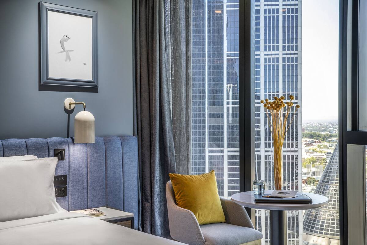 A modern hotel room with a large window, a framed picture on the wall, a bedside table with a lamp, a sofa with a yellow pillow, and a small round table with a vase of dried flowers.