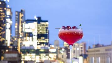 A cocktail glass with a red drink, garnished with berries and a sprig, on a table with a cityscape background.