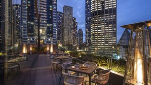 Un área de comedor al aire libre con mesas y sillas, rodeada de edificios altos al anochecer.