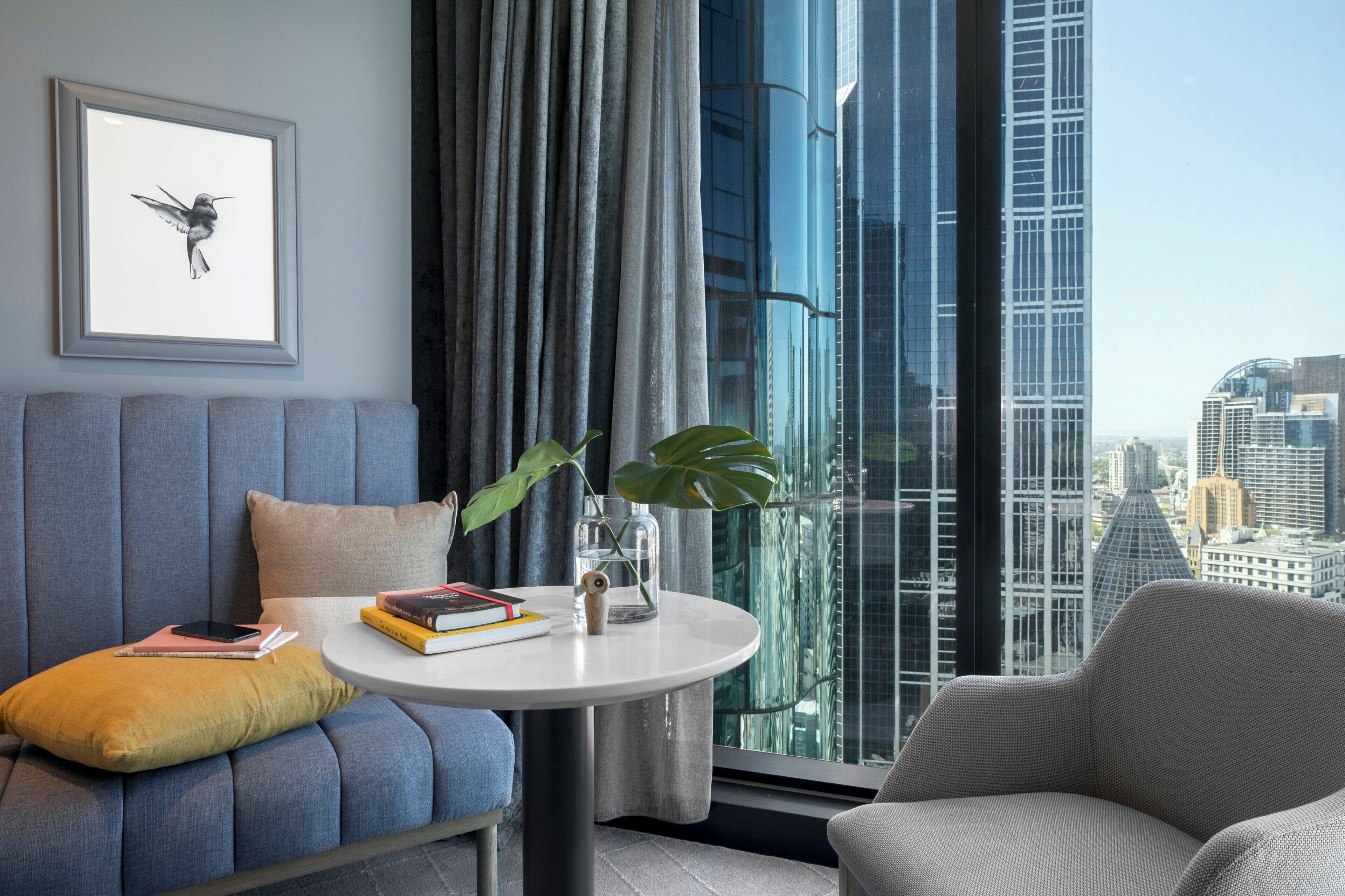 A modern living room with a sofa, a round table, and a view of a cityscape through large windows.
