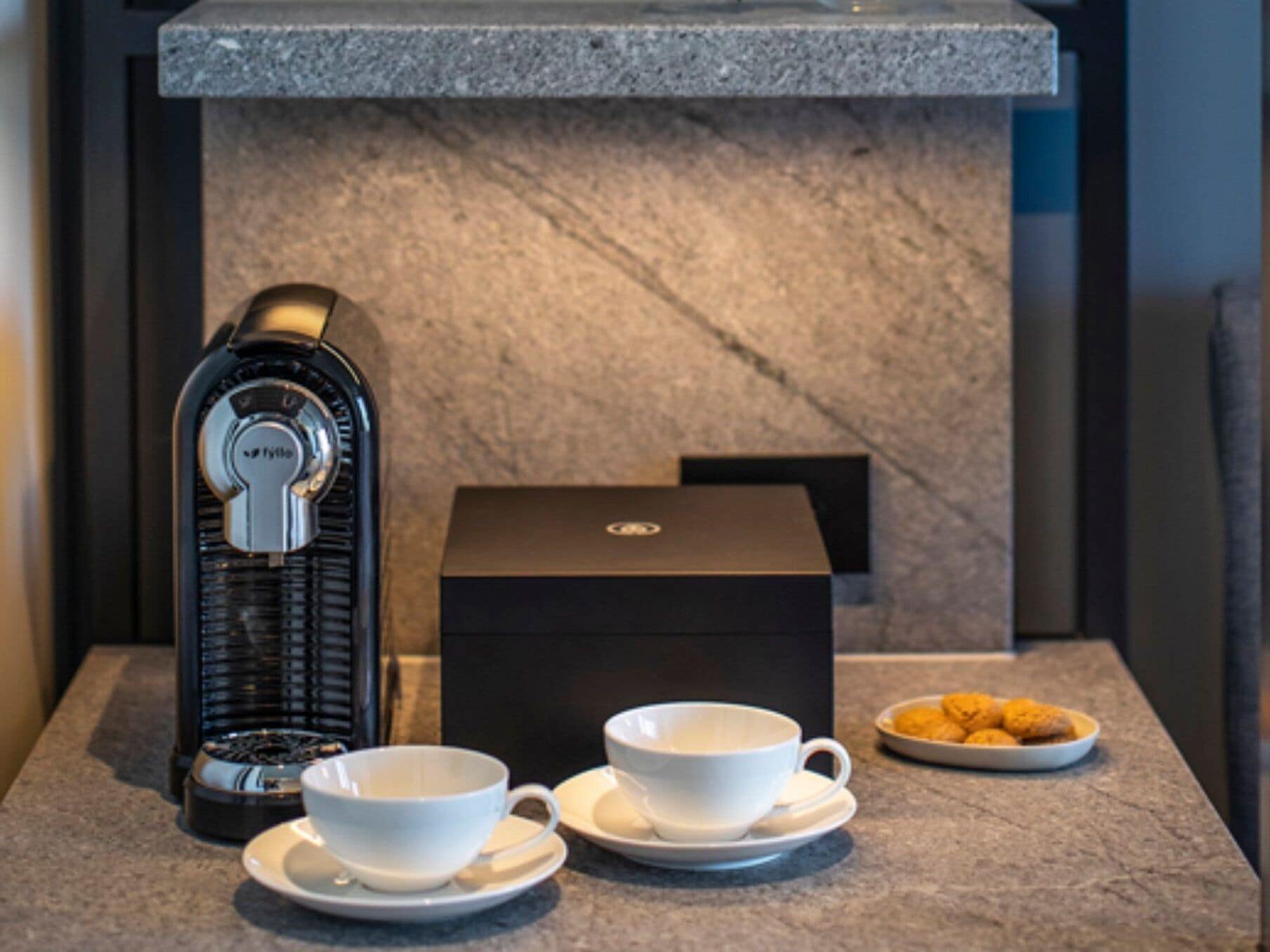 A coffee machine, two white cups on saucers, a plate of cookies, and a black box on a marble countertop.