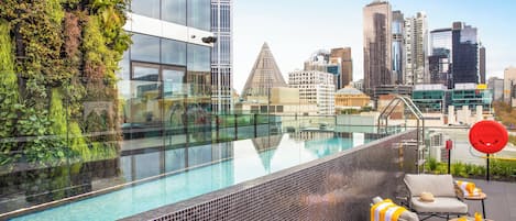 A rooftop pool with a city skyline view, a glass barrier, and outdoor seating.