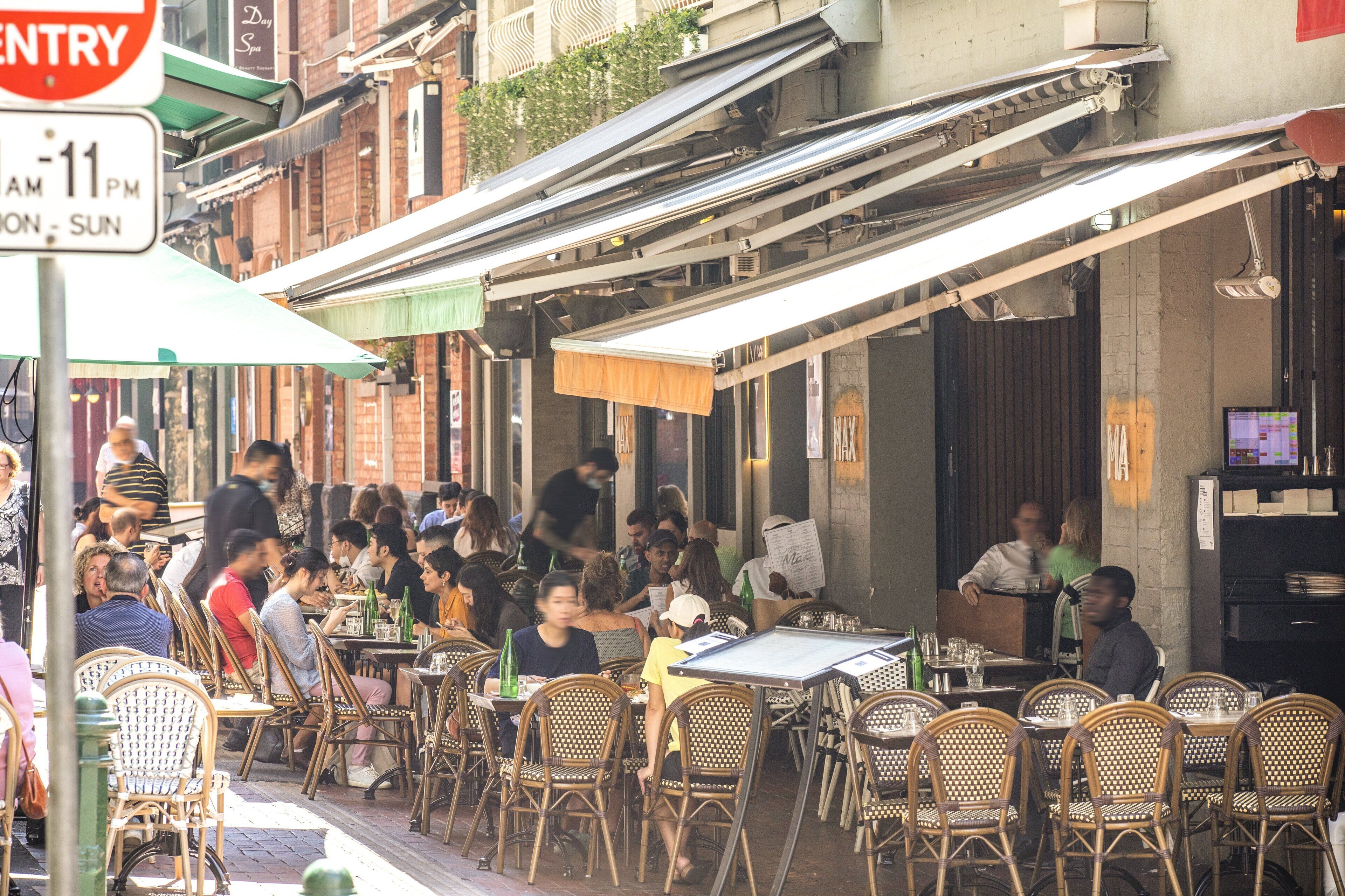 An outdoor cafe with wicker chairs and tables, a covered seating area, and a sign indicating operating hours.