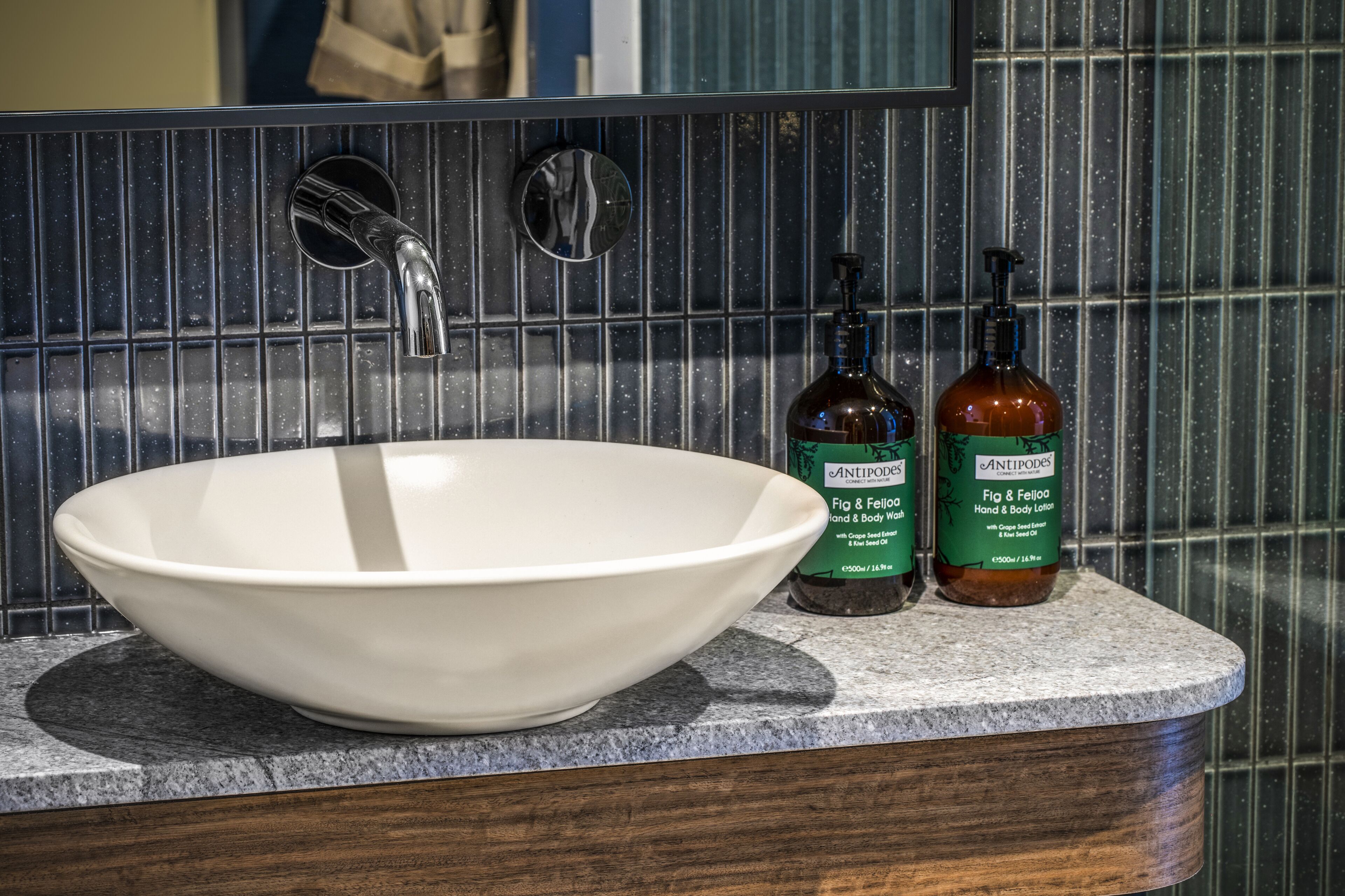 A white ceramic sink with a modern faucet, two bottles of Antipodes hand and body lotion, and a mirror above the sink.