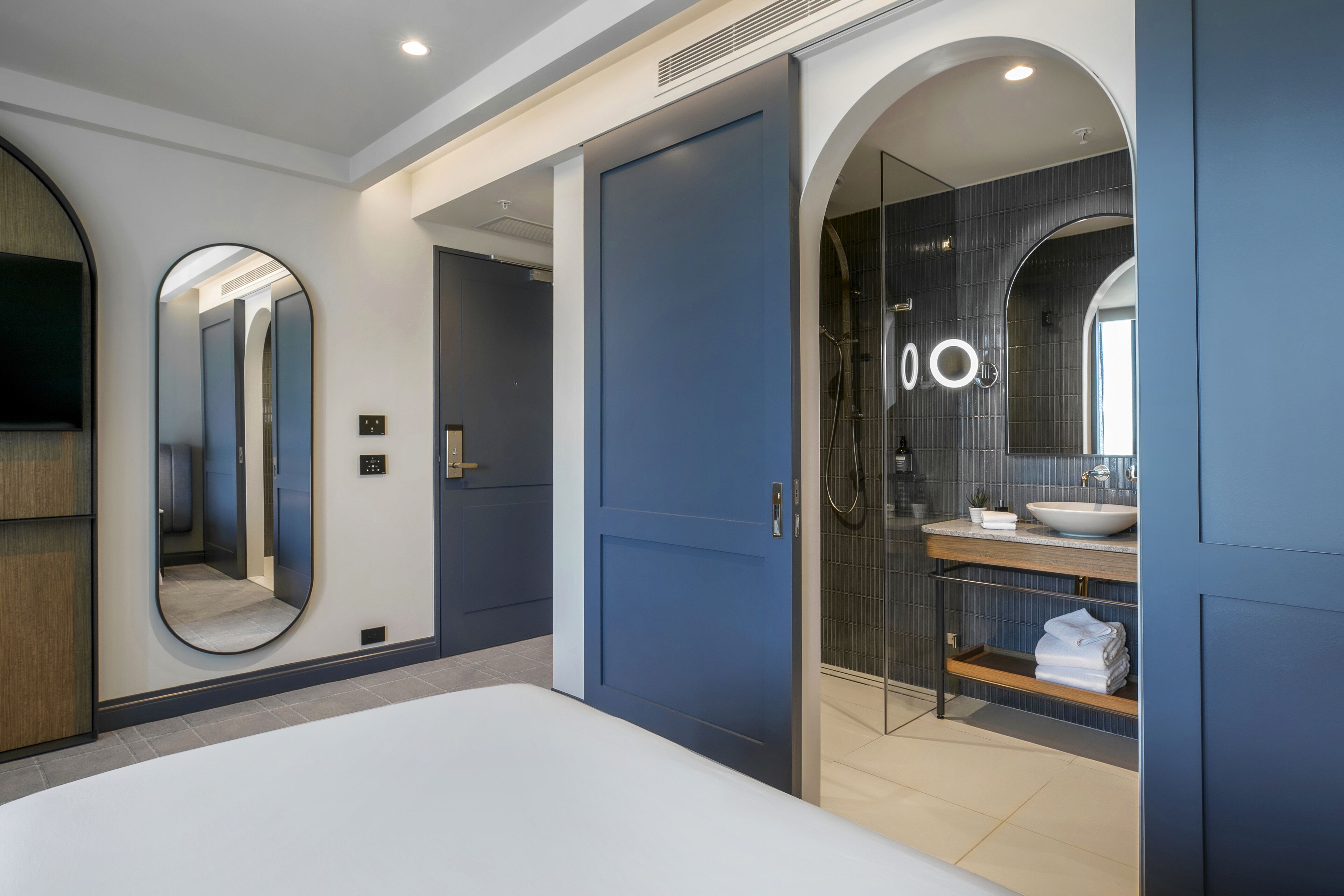 A modern hotel room with a large bed, a walk-in closet, a bathroom with a large mirror, and a shower.