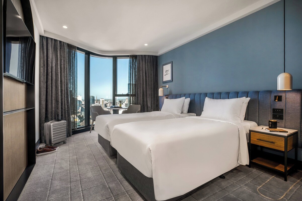 Habitación de hotel con dos camas, un escritorio, una silla, un ventanal con cortinas y vistas a la ciudad.