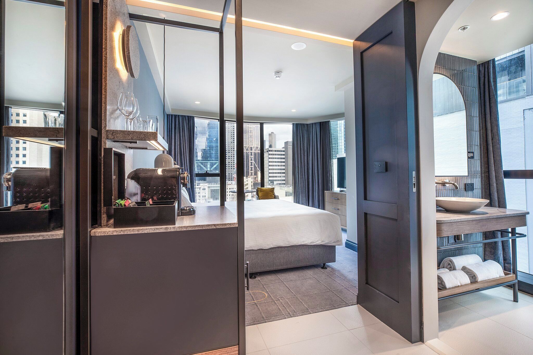 A modern hotel room with a bed, a seating area with a coffee machine, a large window with a city view, and a bathroom with a sink and towel rack.