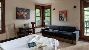 A bedroom with a bed, a dark-colored sofa, a small table with chairs, and two windows with wooden frames.