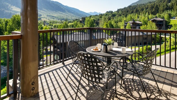 A wooden deck with a round table and chairs, overlooking a mountainous landscape with buildings.