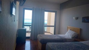 Habitación de hotel con dos camas, ventilador de techo, una mesita y vista al océano desde el balcón.