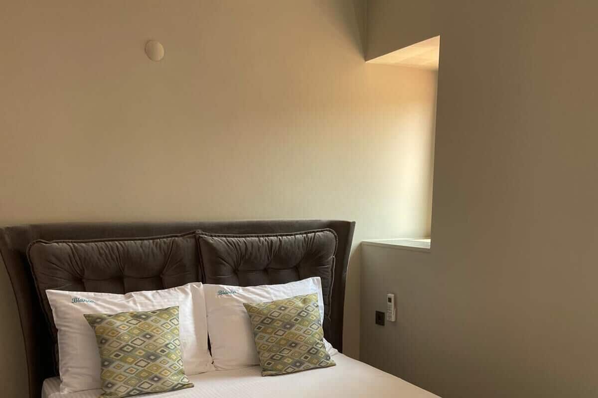 A neatly made bed with a patterned bedspread and two pillows, a tufted headboard, and a small window to the right.