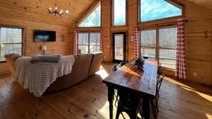 A cozy cabin interior with a wooden ceiling, large windows, a sofa, a dining table, and a TV mounted on the wall.