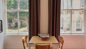 A room with a large window, a wooden table with a bowl of magazines, two wooden chairs, and a view of a building and greenery outside. - St Martins View - Two Bedroom Apartment, Sleeps 4 (York)