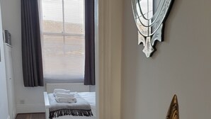 A bedroom with a bed, a bedside table with a Buddha statue, a mirror, and a window with curtains. - St Martins View - Two Bedroom Apartment, Sleeps 4 (York)