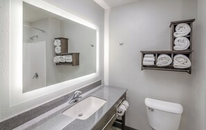 A bathroom with a large mirror, a sink, a toilet, and shelves with towels. - My Place Hotel - Marion, OH (Marion)