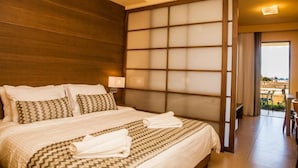 A hotel room with a large bed, a wooden headboard, a sliding glass door leading to a balcony, and a view of the beach. - Ilion Beach Hotel (Platanias)