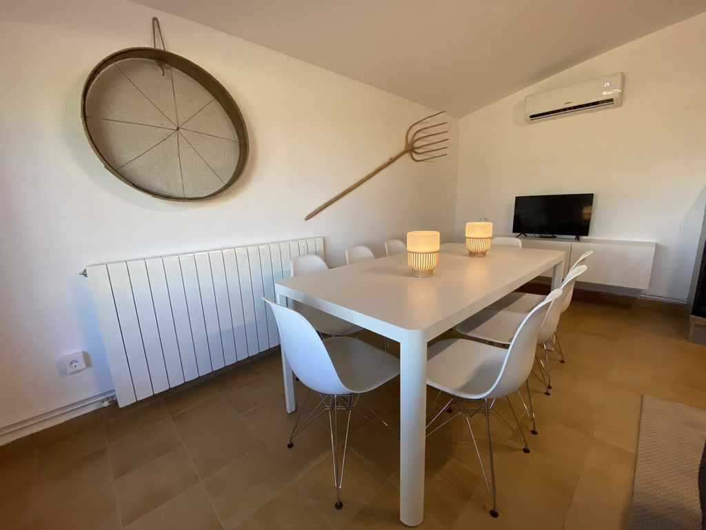 A dining area with a white table, four white chairs, two table lamps, a hanging pot rack, a television, and a wall-mounted air conditioner.