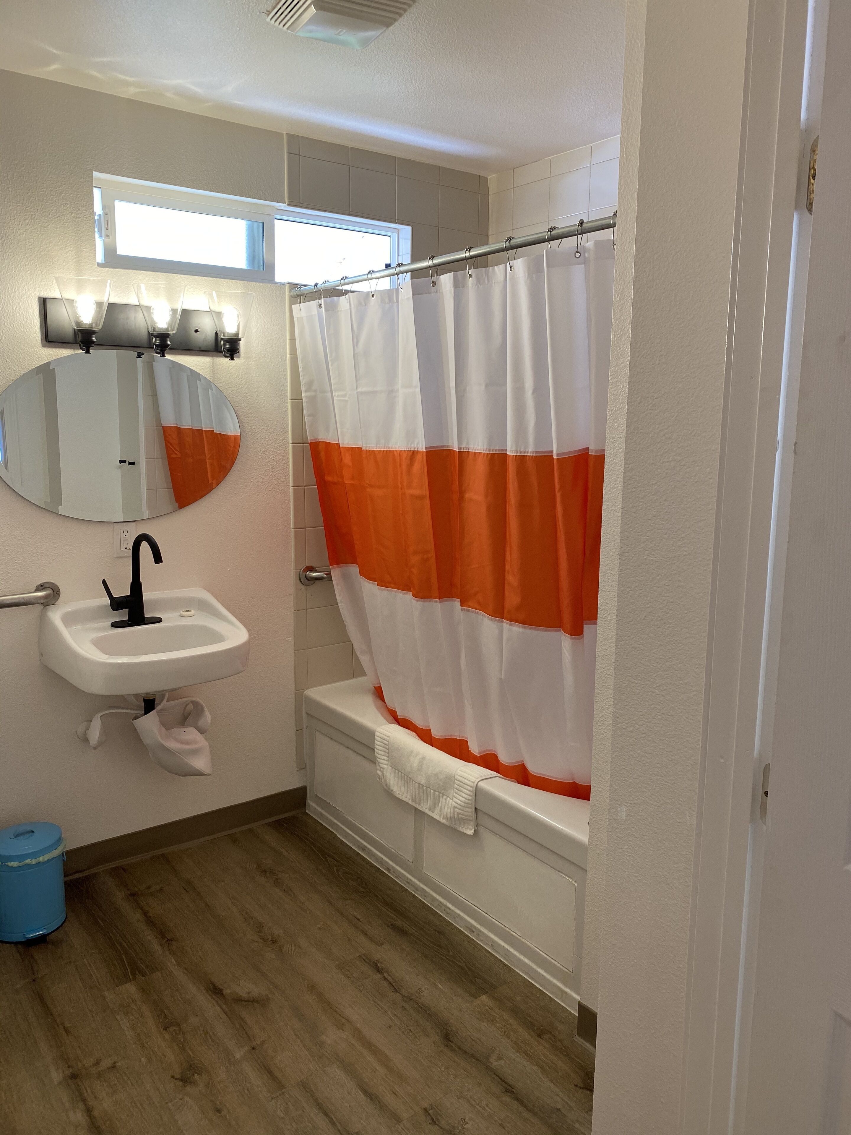 Un baño con lavamanos blanco, un espejo, cortina de ducha y un cubo de basura azul.