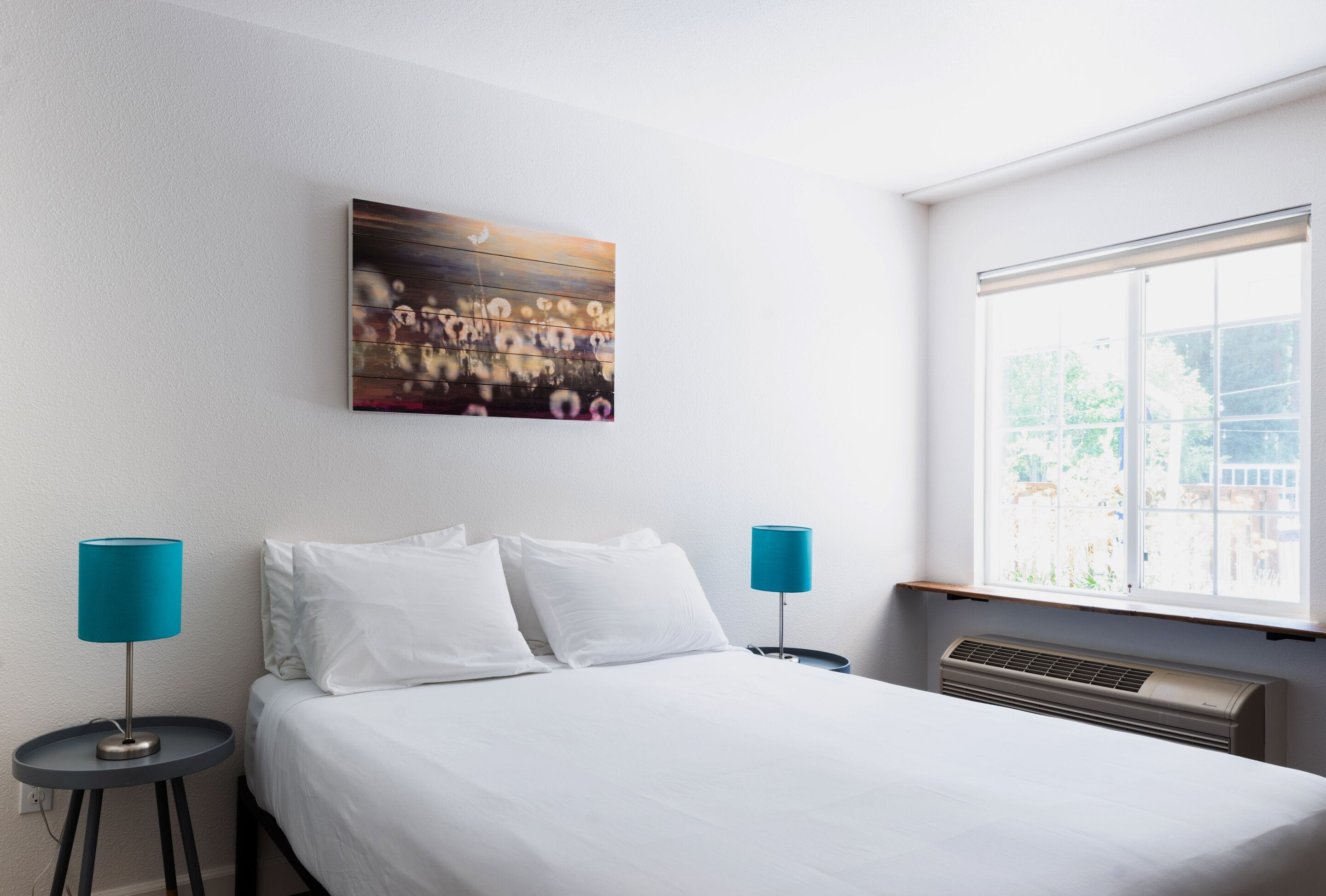 A hotel room with a bed, two bedside tables with lamps, a window with a view, and a painting on the wall.